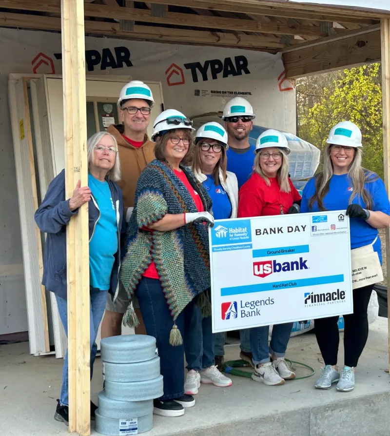 Bank volunteers in hard hats at a Habitat for Humanity build, holding a Bank Day sign
